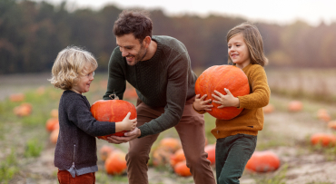 A Dad is at a pumpkin patch with two young children. One child is holding a pumpkin and the Dad is handing off a pumpkin to another child.